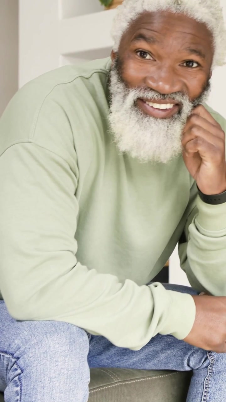 Vertical video: Pausing to speak, senior man leaning on sofa in green top, revealing yellow pillow
