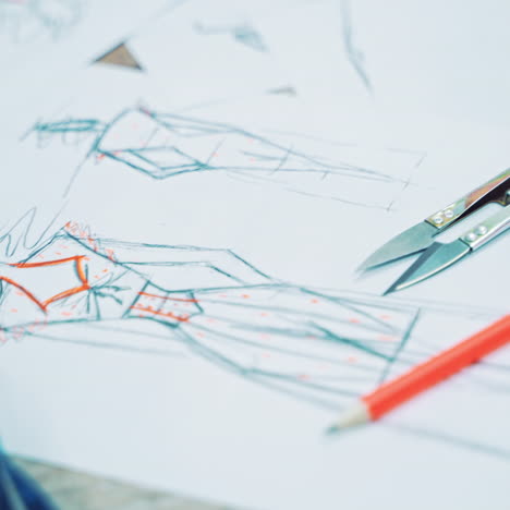 designer drawings of fashionable clothes on papers. tailor's sketches on papers, a pencil and a blue tape measure on the wooden table indoors. camera moves left.
