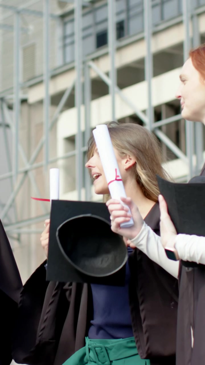 Vertikales Video: Zwei Absolventen in schwarzen Talaren wenden sich der Kamera zu und halten Diplome und Absolventenhüte auf dem Campus hoch.