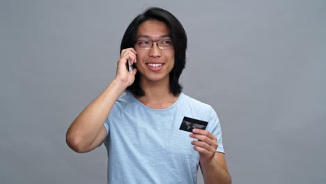 handsome cheerful young asian man holding credit card and talking by phone