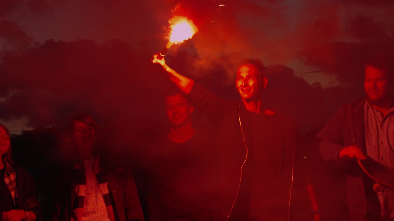 un grupo de adolescentes felices con una luz de señal en la mano y agitando caminando hacia adelante hacia la cámara por la noche.