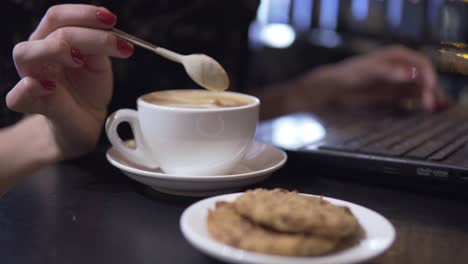 una mujer mezclando su capuchino usando una cuchara y trabajando con una computadora portátil en el fondo.