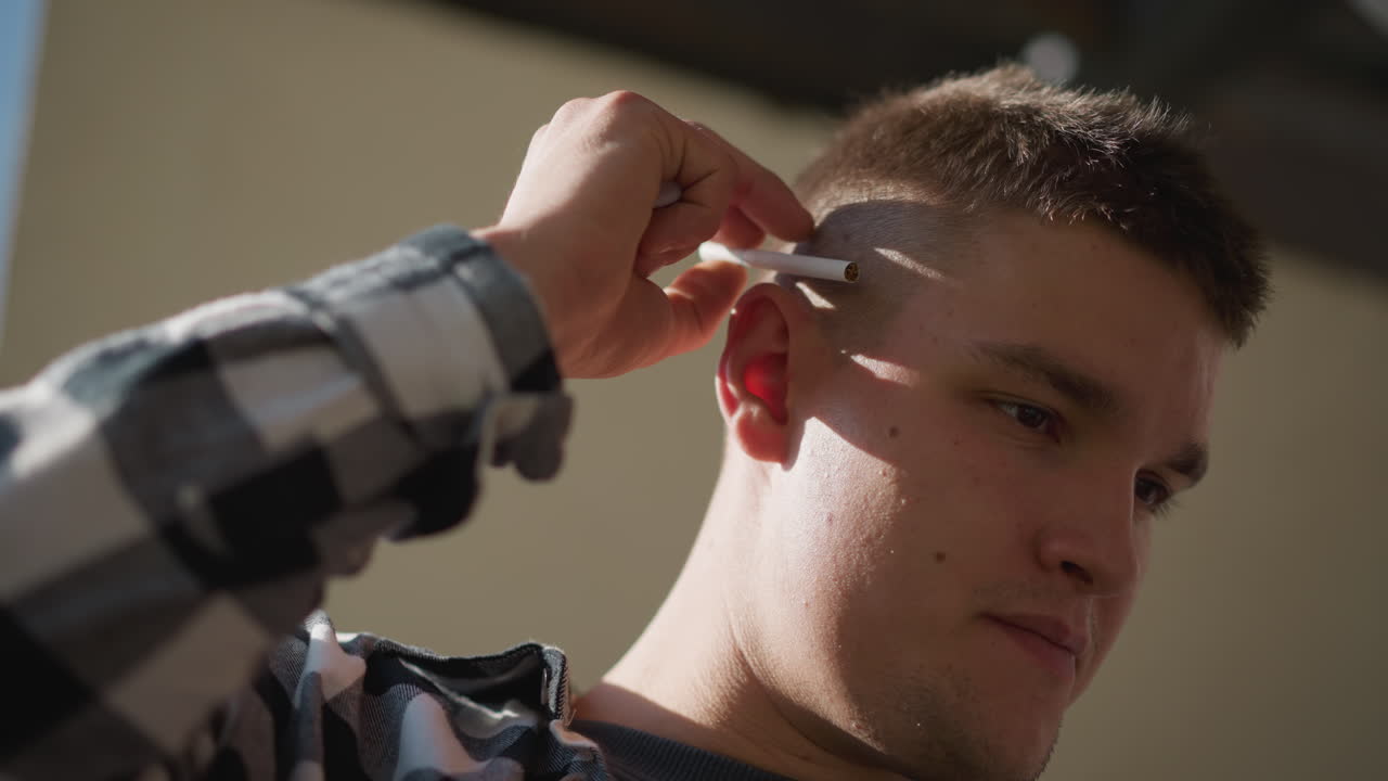 close up of young man in black white plaid shirt on metal staircase retrieving cigarette from ear raises hand to mouth preparing to light tip with lighter against blurred sky backdrop