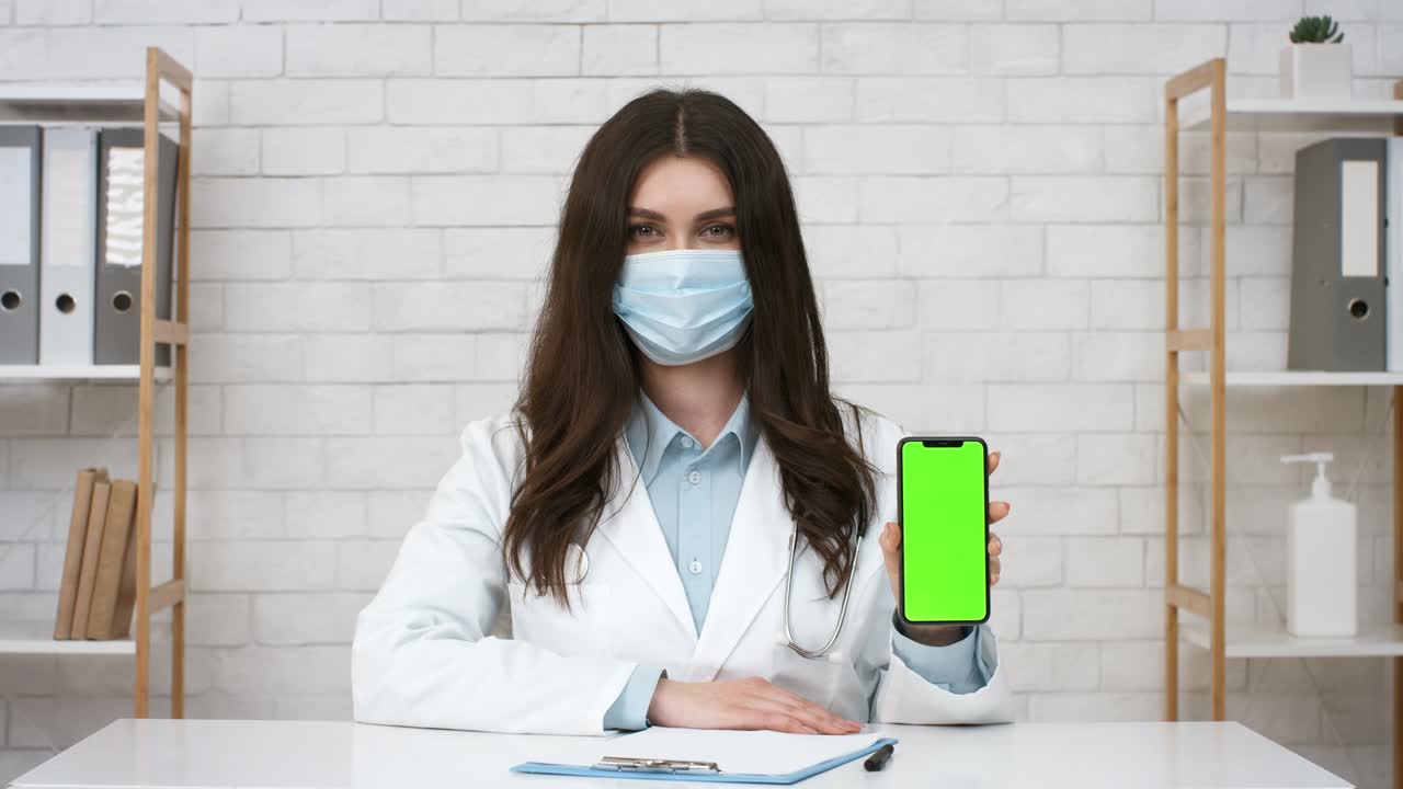 médico femenino mostrando la pantalla del teléfono celular usando una máscara facial en la oficina