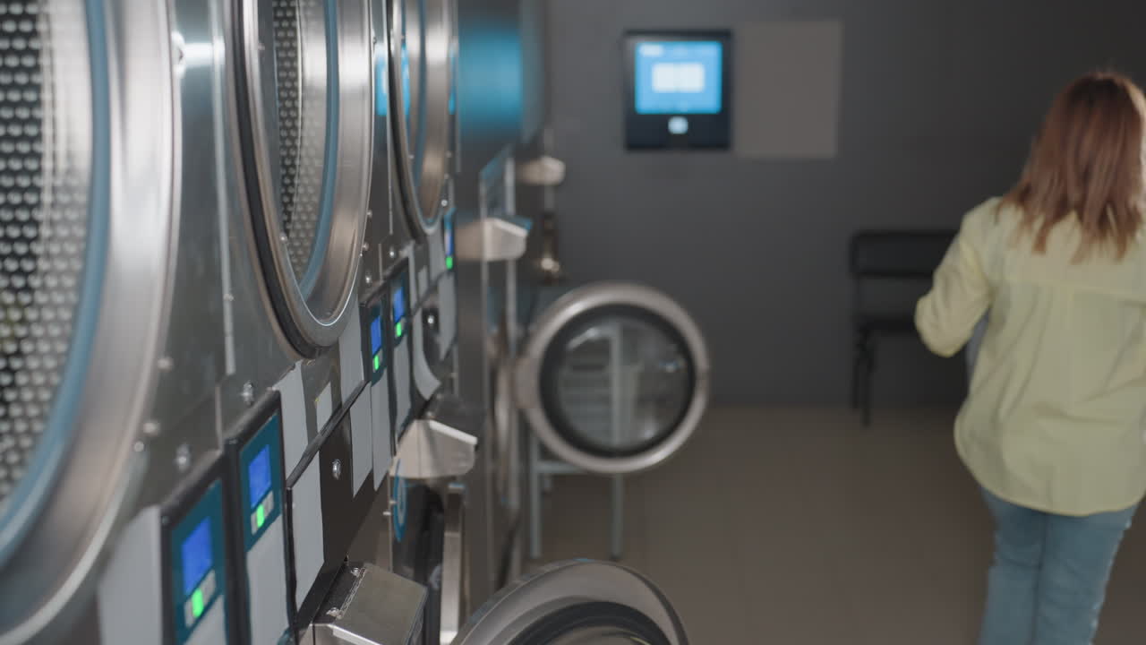 High angle view of young woman with laundry basket walking away from industrial washer, door open, stainless machines in row, laundromat interior, routine workflow, cleaning operation in progress