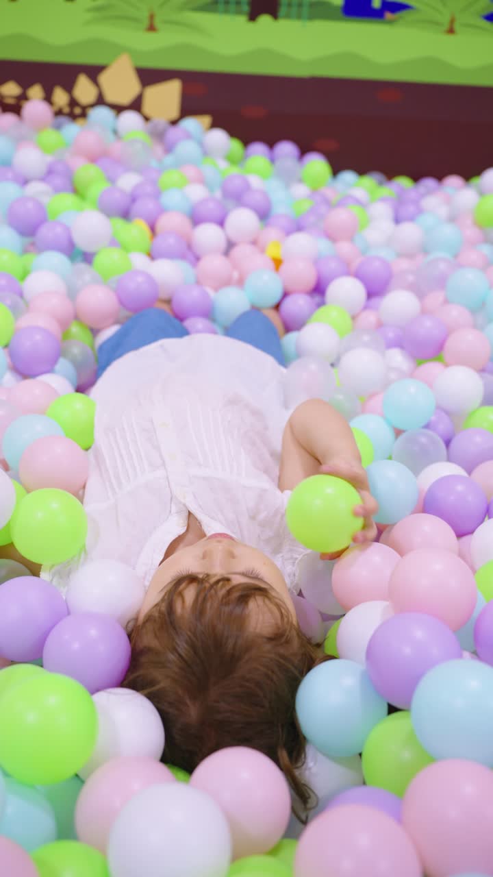 A child lies comfortably amidst a sea of colorful pastel balls and throwing balls in the air on a playground, throwing them