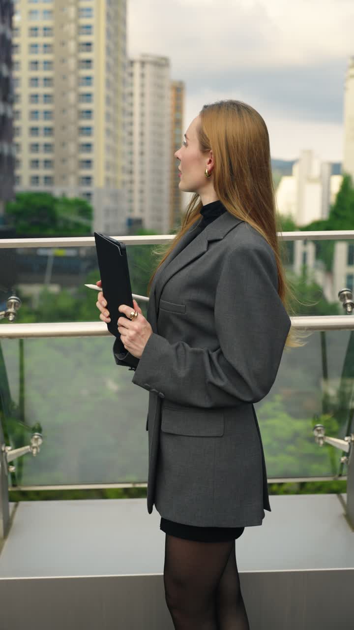 Confident businesswoman stands on a terrace, reviewing a digital tablet and making notes with a stylus, surrounded by city skyscrapers and modern architecture, nodding for success