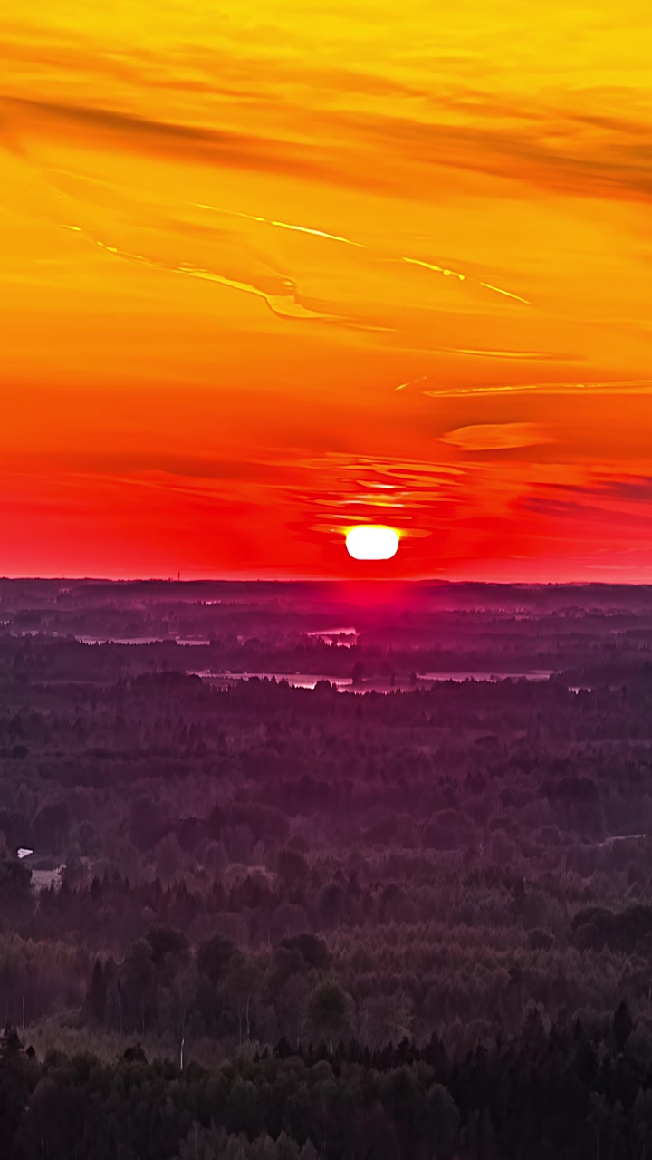 Rekaman timelapse vertikal dari udara saat matahari terbenam di atas cakrawala datar, menerangi langit dengan nuansa merah dan oranye cerah di atas hutan dan danau berbayang gelap di pedesaan Latvia.