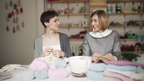 Knitting-woman-smiling-and-talking-with-friend-sitting-at-table-in-workshop