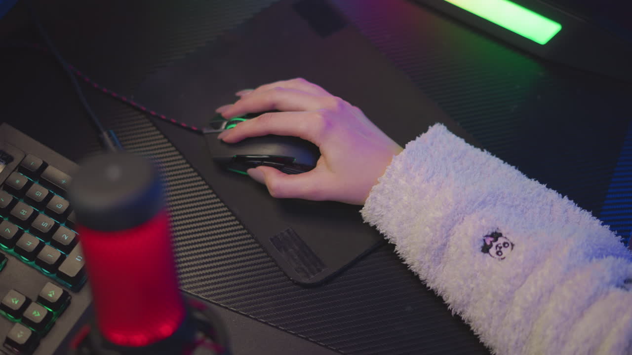 Aerial view of hand with polished nail in fluffy sweater using wired gaming mouse on black mousepad, surrounded by subtle ambient RGB lighting from keyboard and LED strip
