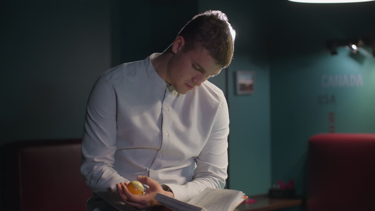 Man in white shirt sits on green pool table, reading book while rolling billiard ball in hand. Pool balls scattered across table, creating focused, moody atmosphere as camera zooms in for close-up shot