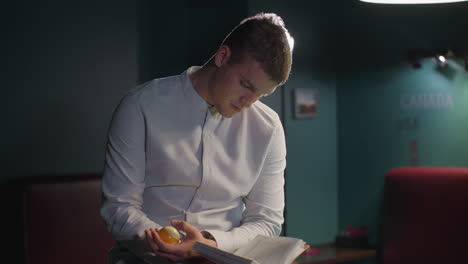 hombre de camisa blanca se sienta en la mesa de billar verde, leyendo un libro mientras gira la pelota de billar en la mano. bolas de billar esparcidas por la mesa, creando una atmósfera enfocada y de mal humor mientras la cámara hace zoom para una toma de primer plano