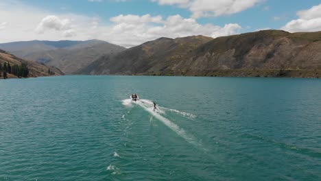 SLOWMO---Waterskiing-behind-boat-on-Lake-Dunstan-near-Clyde-dam,-Central-Otago,-New-Zealand-with-mountains-and-clouds-in-background