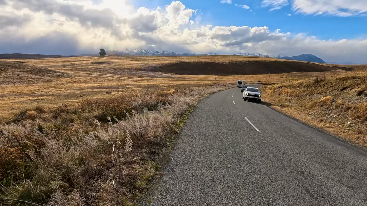 A camper van travels along a winding rural road through golden autumn fields under dramatic skies near Lake Tekapo, captured in smooth tracking shots