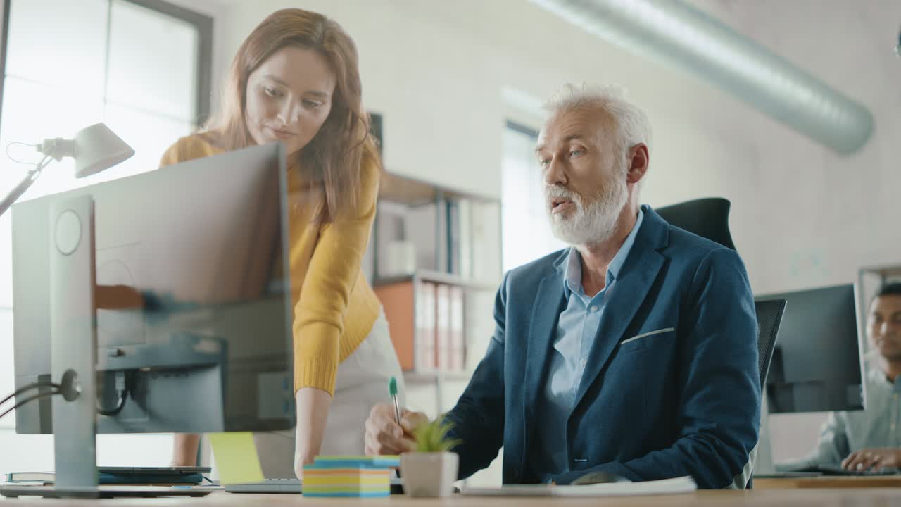 Handsome Middle Aged Creative Engineer Sitting at the Desk working on Desktop Computer Has Discussion with Beautiful Female Project Manager. Office with Friendly Colleagues Chatting, Finding Solutions