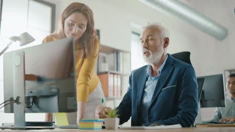 handsome middle aged creative engineer sitting at the desk working on desktop computer has discussion with beautiful female project manager. office with friendly colleagues chatting, finding solutions