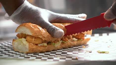 chef uses red serrated knife to cut fried shrimp poboy sandwich in half, slow motion close up 4k