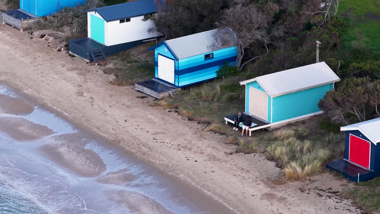 Drone glides above pastel beach huts and sandy shoreline in soft daylight, steady aerial perspective