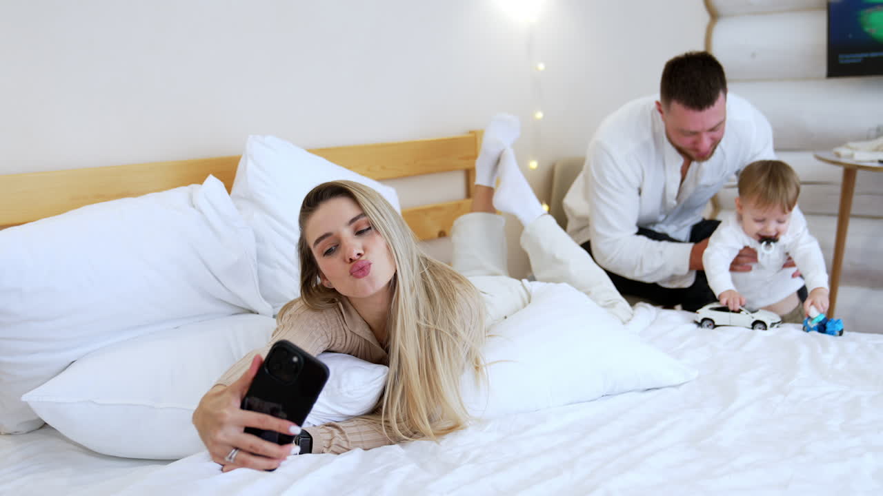 Beautiful Caucasian blonde woman lying on the bed taking selfie. Man and a baby boy play with toys at backdrop.