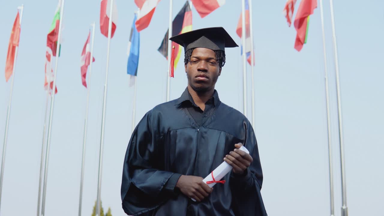 Young african american male graduate standing in front of the camera with a diploma and books in her hands. The student stands outside with the international flags on background.