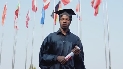 joven graduado afroamericano de sexo masculino de pie frente a la cámara con un diploma y libros en sus manos. la estudiante está de pie afuera con las banderas internacionales en el fondo.