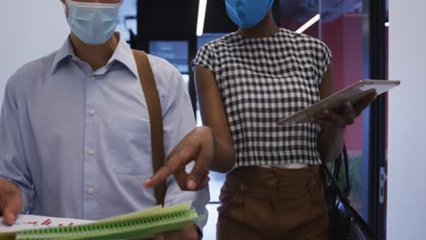 diverse business colleagues wearing face masks using tablet in discussion walking through office