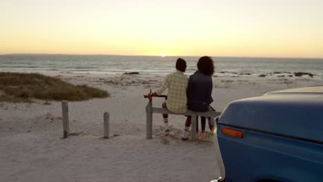 Rear-view-of-African-american-couple-sitting-together-on-bollard-at-beach-4k