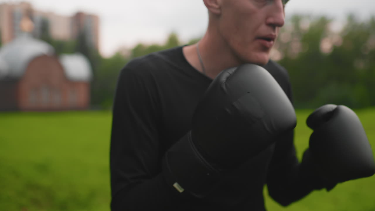 Treino individual de shadowboxing urbano em parque, com a guarda alta e footwork preciso, luvas pretas, prédios distantes no horizonte, gramado verde ao redor, respiração focada e movimento repetitivo, defensivo