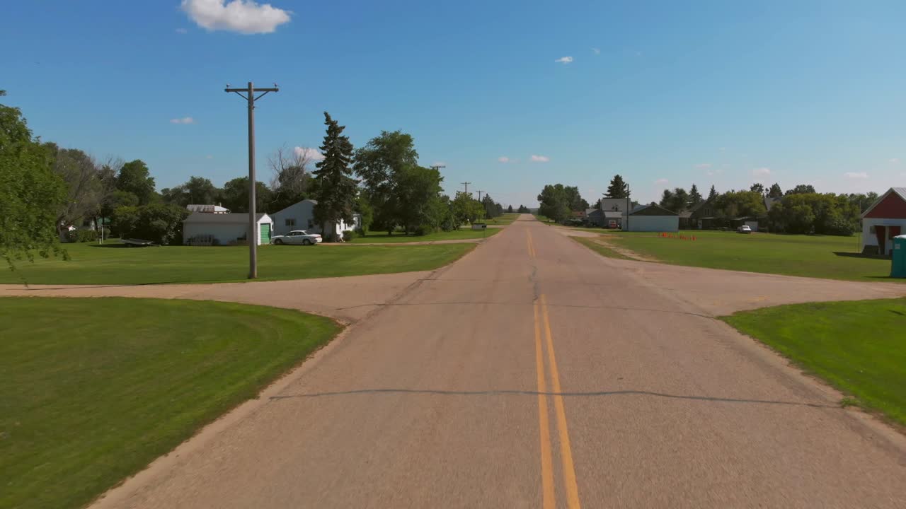 Low aerial shot flying backward along a quiet rural road lined with small houses, trees, and open lawns under a bright blue summer sky