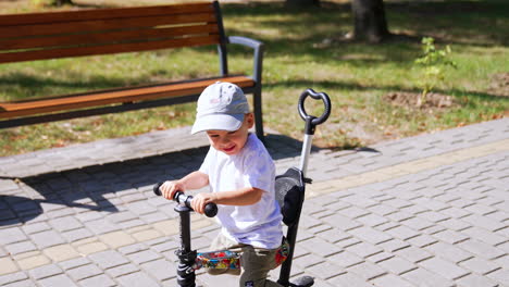 little cheerful caucasian boy in a cap riding his bike. cute toddler pushes from ground with his legs. positive kid in the park on sunny summer day.