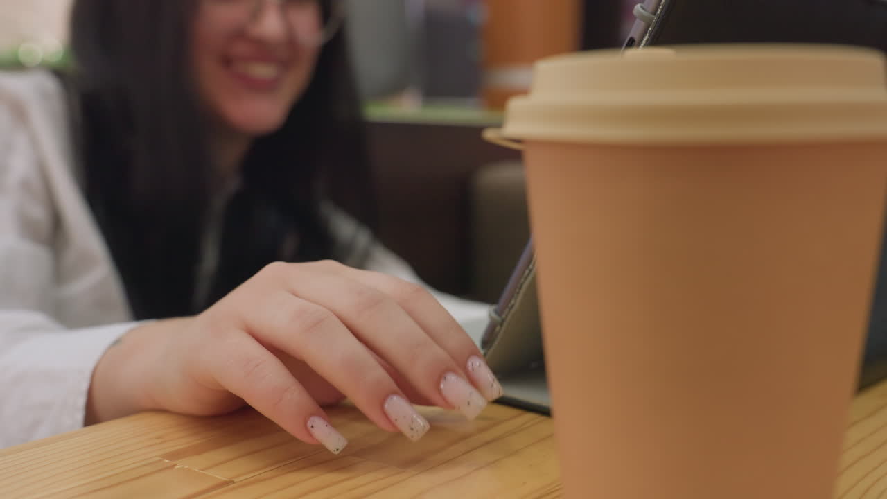 close up of takeaway coffee cup on wooden table with woman manicured hand tapping gently as she smiles in background creating cozy ambient mood in indoor setting with digital tablet beside