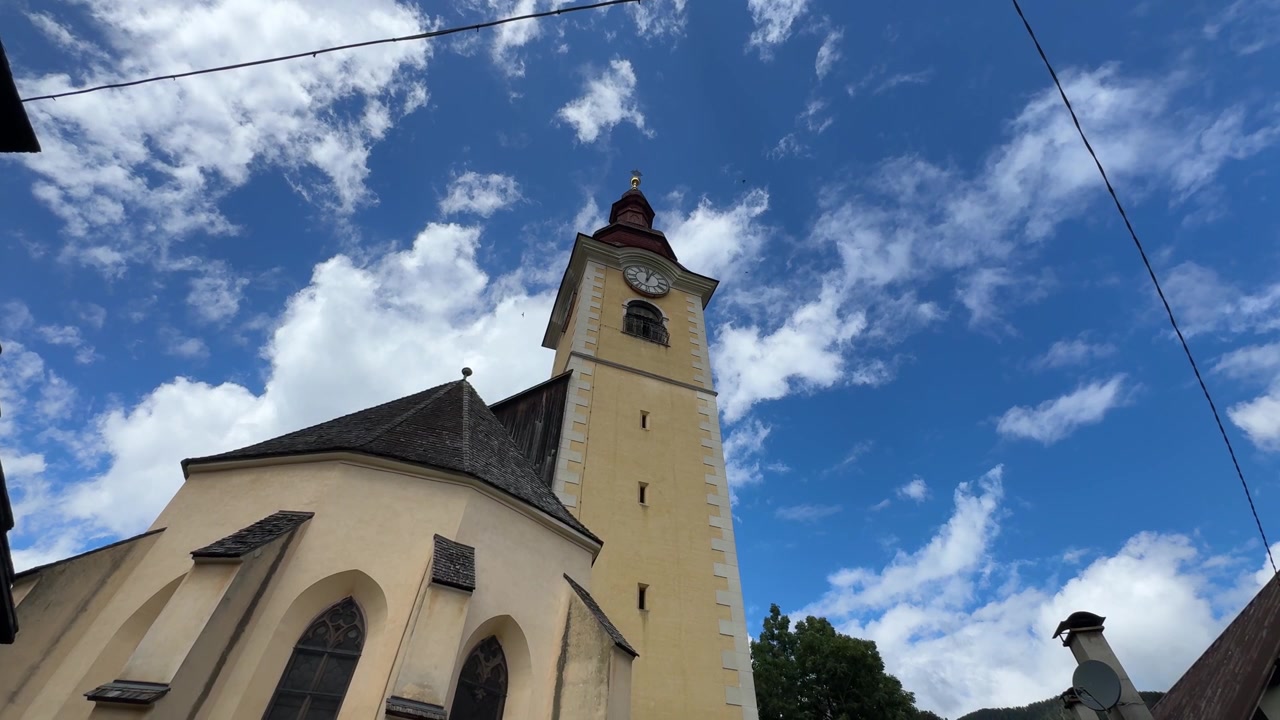Vista em ângulo baixo de uma torre sineira amarela histórica com cúpula em forma de cebola em Tarvisio, Itália, emoldurada contra um céu azul vibrante com nuvens fofas, destacando a herança alpina