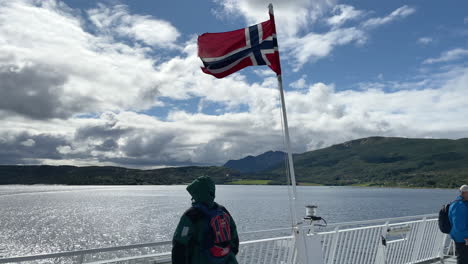 Medium-static-shot-of-a-person-looking-out-over-the-railing-of-a-ferry-boat-with-a-Norwegian-flag-flying-and-a-partially-cloudy-sky-and-green-mountains-in-the-background