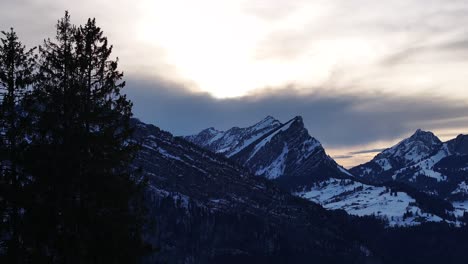 Toma-Reveladora-De-Los-Alpes-Suizos-Durante-La-Puesta-De-Sol,-Con-Siluetas-De-Pinos-En-Primer-Plano-Y-Picos-Nevados-Al-Fondo-Bajo-Un-Cielo-Espectacular.