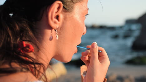 Closeup-shot-of-a-beautiful-woman-with-a-natural-look-eating-with-spoon-an-ice-cream-on-the-beach-while-the-waves-are-breaking-around-and-the-sun-is-illuminating-her-face-and-she-enjoy-the-sea-view