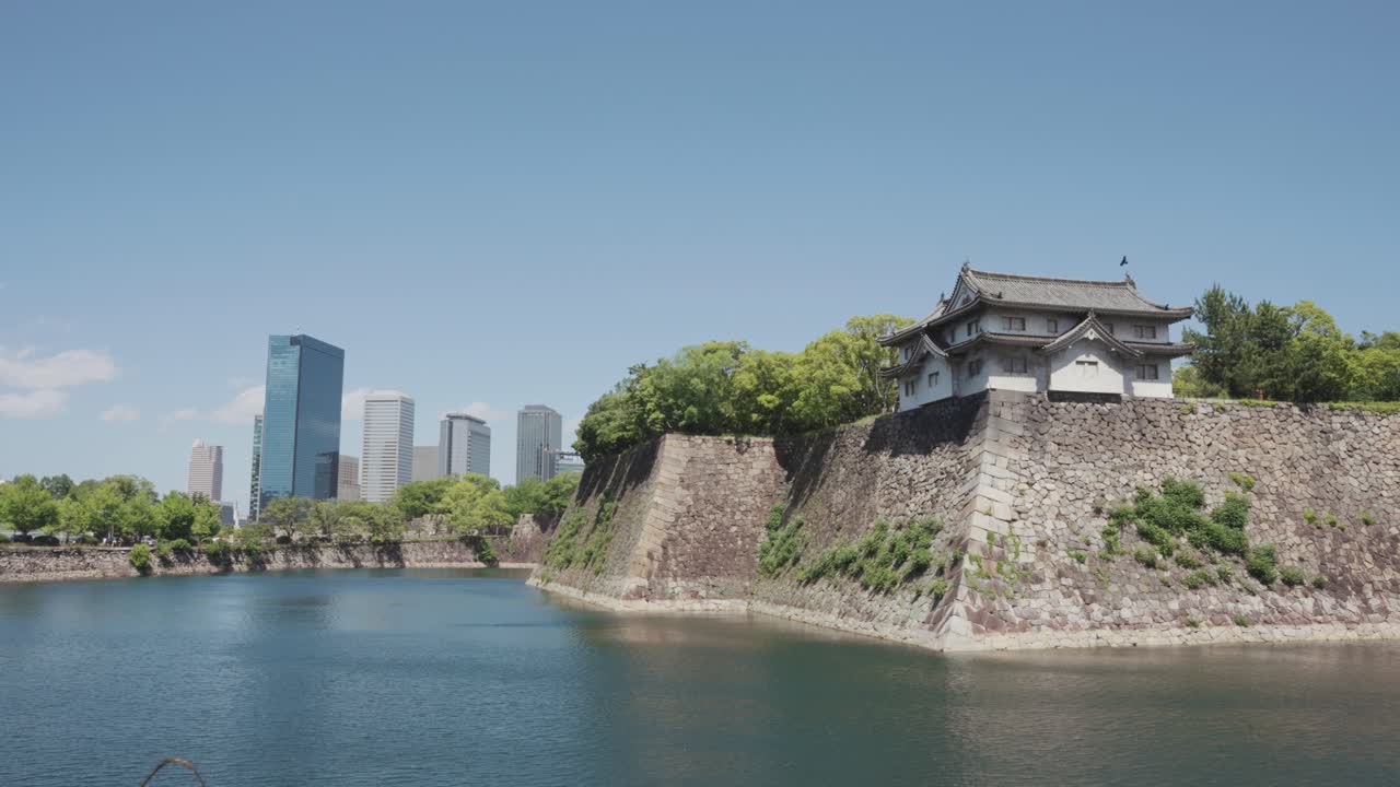 A stunning view of Osaka Castle and its moat, surrounded by lush greenery, with modern skyscrapers in the background under a bright blue sky, blending history and urban scenery.