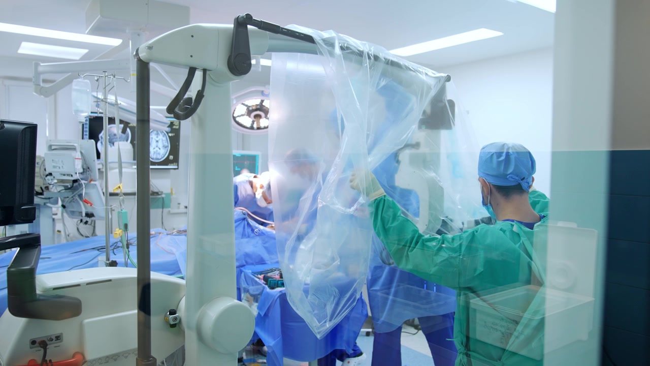 Male medics cover the microscope on the stand with plastic for sterility in the surgery room. Neurosurgeons work at operating table at backdrop.