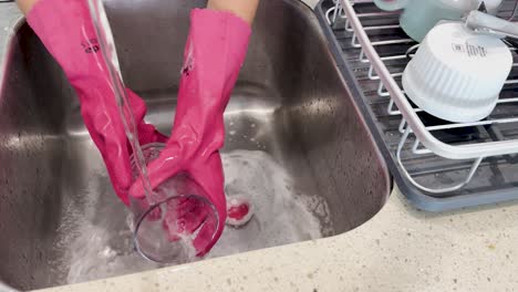 person wearing pink rubber gloves scrubs and rinses a glass under running water in a stainless steel kitchen sink with bright overhead lighting