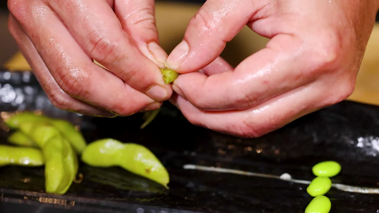 Close-up of chef hands shelling edamame beans on black plate under warm kitchen lighting