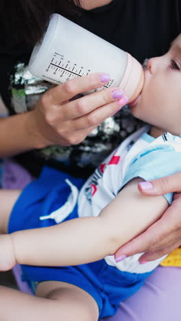 Lovely cute boy eating from bottle and touching socks on his foot. Mommy feeding baby outdoors. Close up. Vertical video