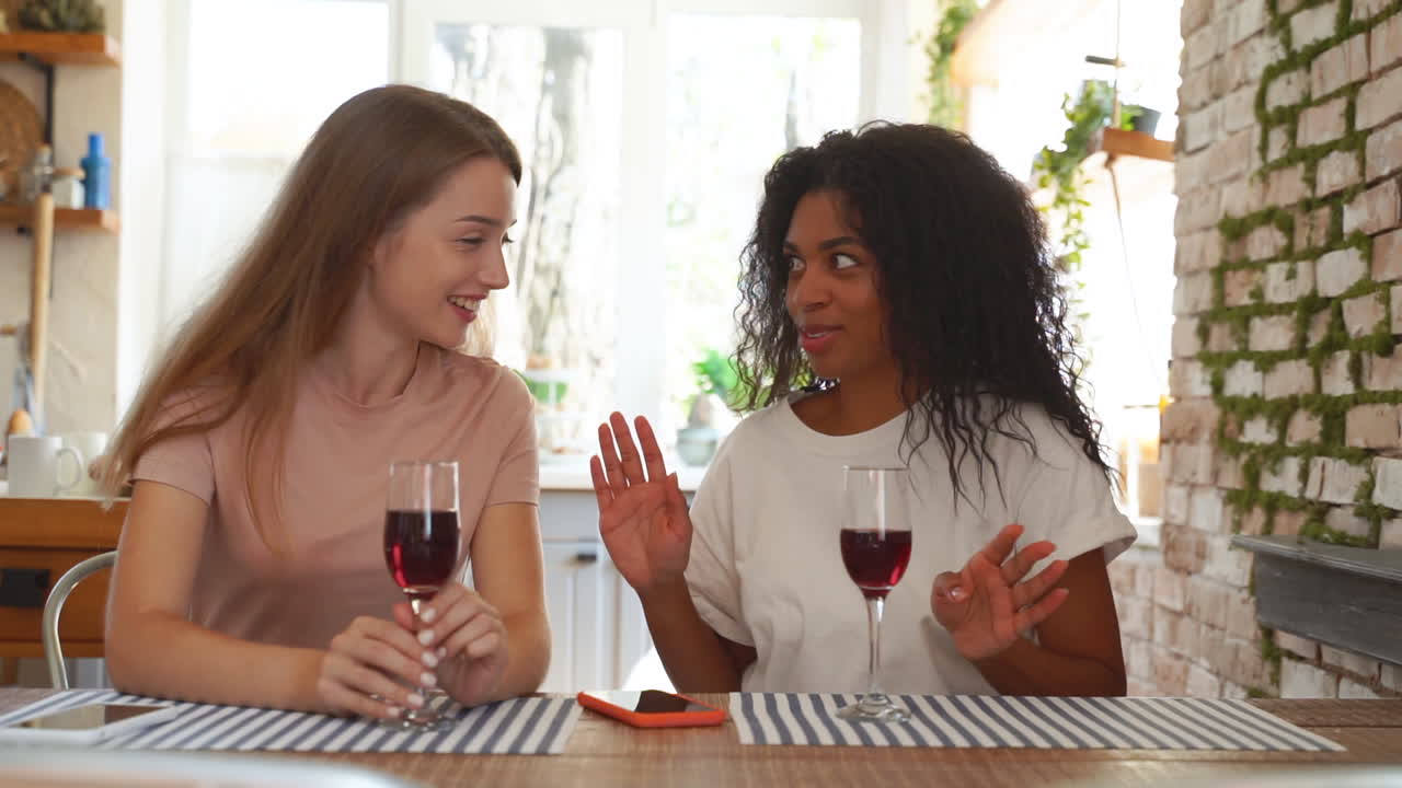 dos amigas, una cuenta secretos al oído, una chica negra y una joven caucásica beben una copa de vino.