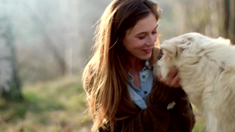 smiling happy woman gently stroking white dog around fire place.real friends people outdoor camping tent vacation in autumn trip.fall sunny day in nature, animal friendship. 4k video