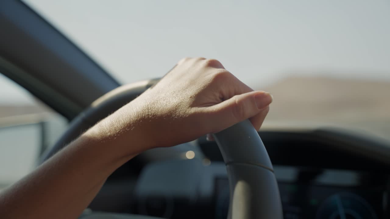 Close up on female hand on steering wheel driving on desert highway with one hand on wheel. Relaxed commute going to work or on vacation. Escaping or going away in non urban road during summer hot day