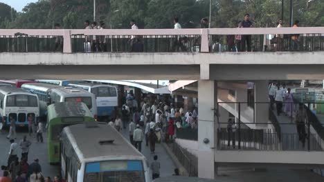 people gather in a bus station getting on and off buses and crossing an overhead walkway