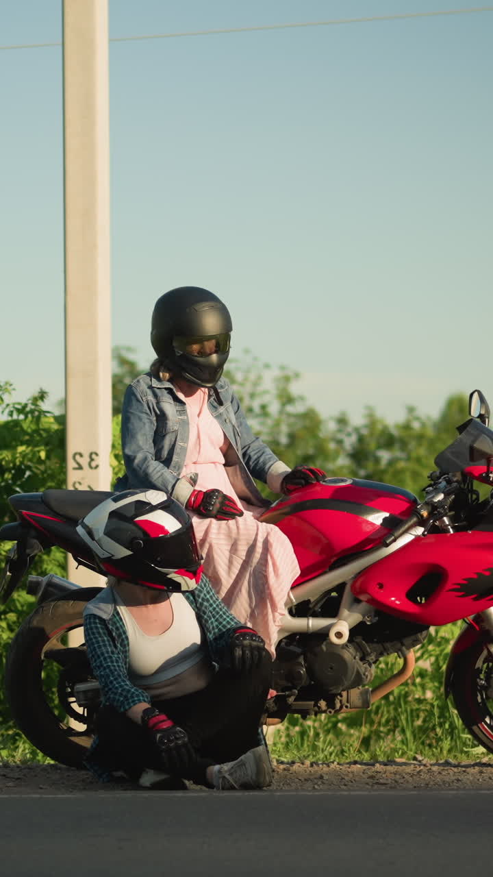 길가에 두 명의 여성, 하나는 빨간 모터사이클에 앉아 왼쪽과 오른쪽을 바라보고 다른 하나는 바닥에 앉았고, 둘 다 같은 방향을 바라고 있습니다.