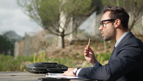 thoughtful business man in suit making notes on papers while eating snacks sitting in chair in a park