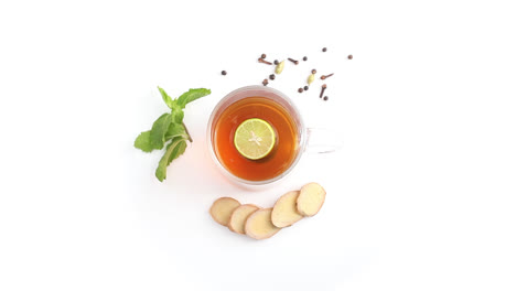 Té-De-Limón-En-Una-Taza-De-Vidrio-Transparente-Con-Clavos-De-Limón-Enteros-Y-En-Rodajas-Cardamomo-De-Pimienta-Negra-En-Fondo-Blanco-Girando-La-Parte-Superior