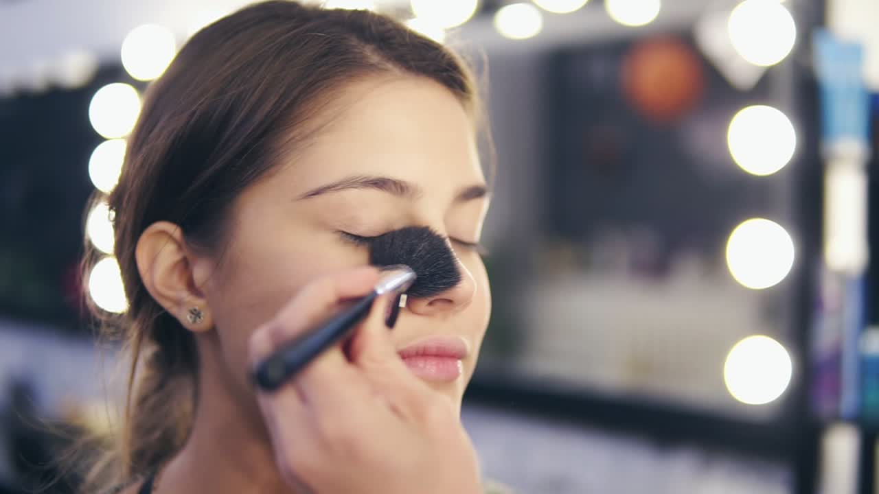 Close Up view of female hands applying facial powder on young woman's skin using special brush. Slow Motion shot