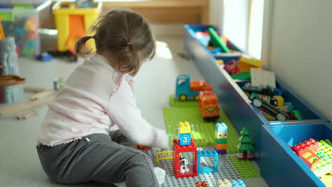 cute funny little girl playing with construction toy blocks at home sitting on a floor by the window