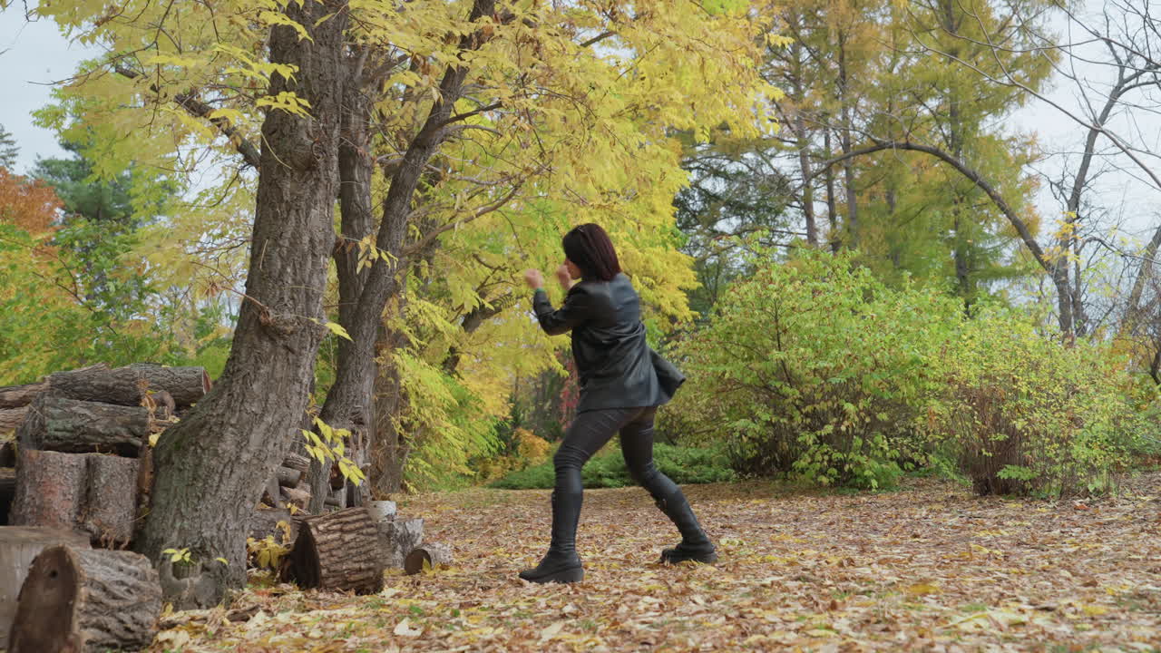 Woman in forest stacking logs, Woman wearing leather jacket lifting wood in forest setting carefully, Woman in rustic boots preparing woodpile amidst leafcovered ground under trees confidently